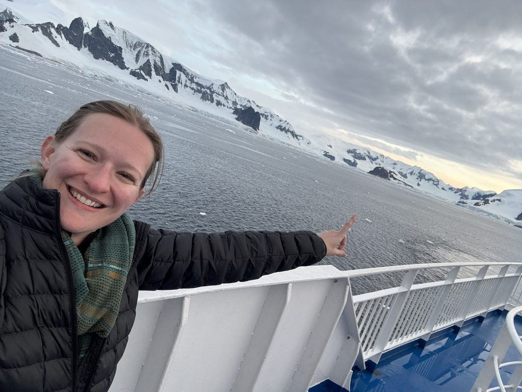 The author, Kaylyn K., smiling and pointing at a snow covered Cuverville Island while visiting Antarctica in February 2025 from the back deck of her expedition ship.