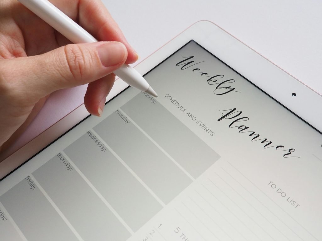 Image of a tablet open to a Weekly Planner page with a hand holding a stylus. The image represents part of the process of working with a personal travel planner. Photo by Jess Bailey Designs