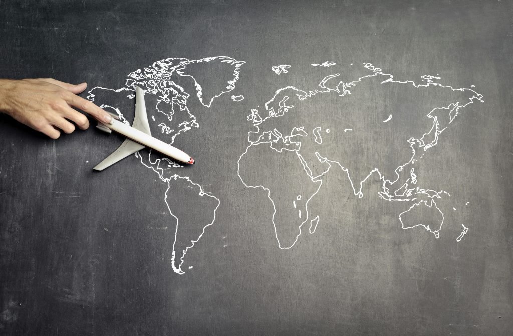 A hand pushes a toy plane, while daydreaming about trip planning, across an outline of a world map on a chalkboard. Stock photo by Andrea Piacquadio