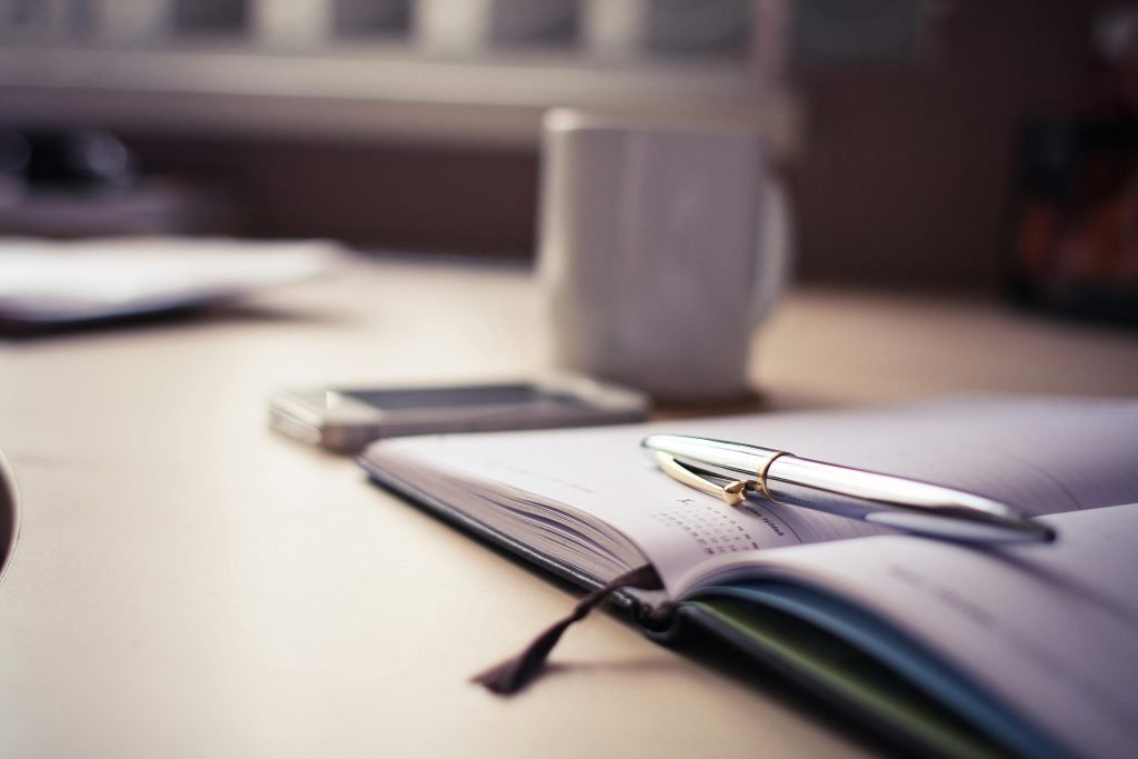 Image of an open book with a pen resting on it and a coffee mug in the background. The image represents the process of working with a personal travel planner. Photo by picjumbo.com