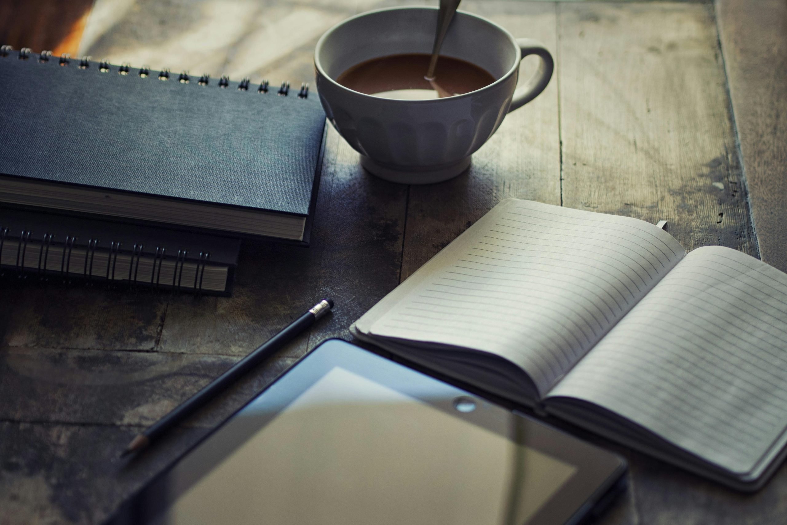 Image of an open journal, coffee cup, iPad, and closed notebook on a table. The image is meant to represent the process of planning a custom travel itinerary.